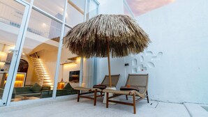 A modern outdoor seating area with a thatched umbrella, wicker chairs, and a glass wall reflecting interior lighting. - Villa Casa Zaya B (Canggu)