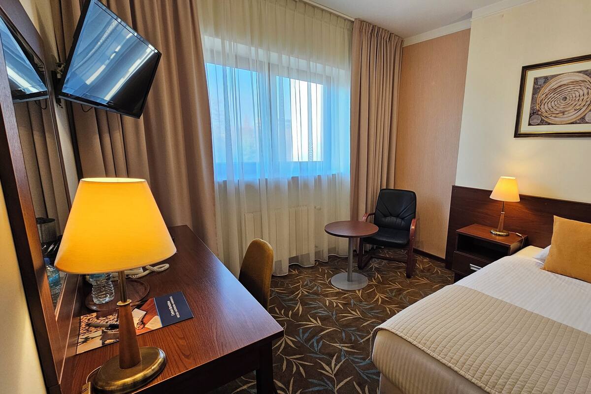 Hotellihuone, jossa on sänky, työpöytä lampun kanssa, tuoli, pieni pöytä, televisio ja seinällä oleva kehystetty kuva.
