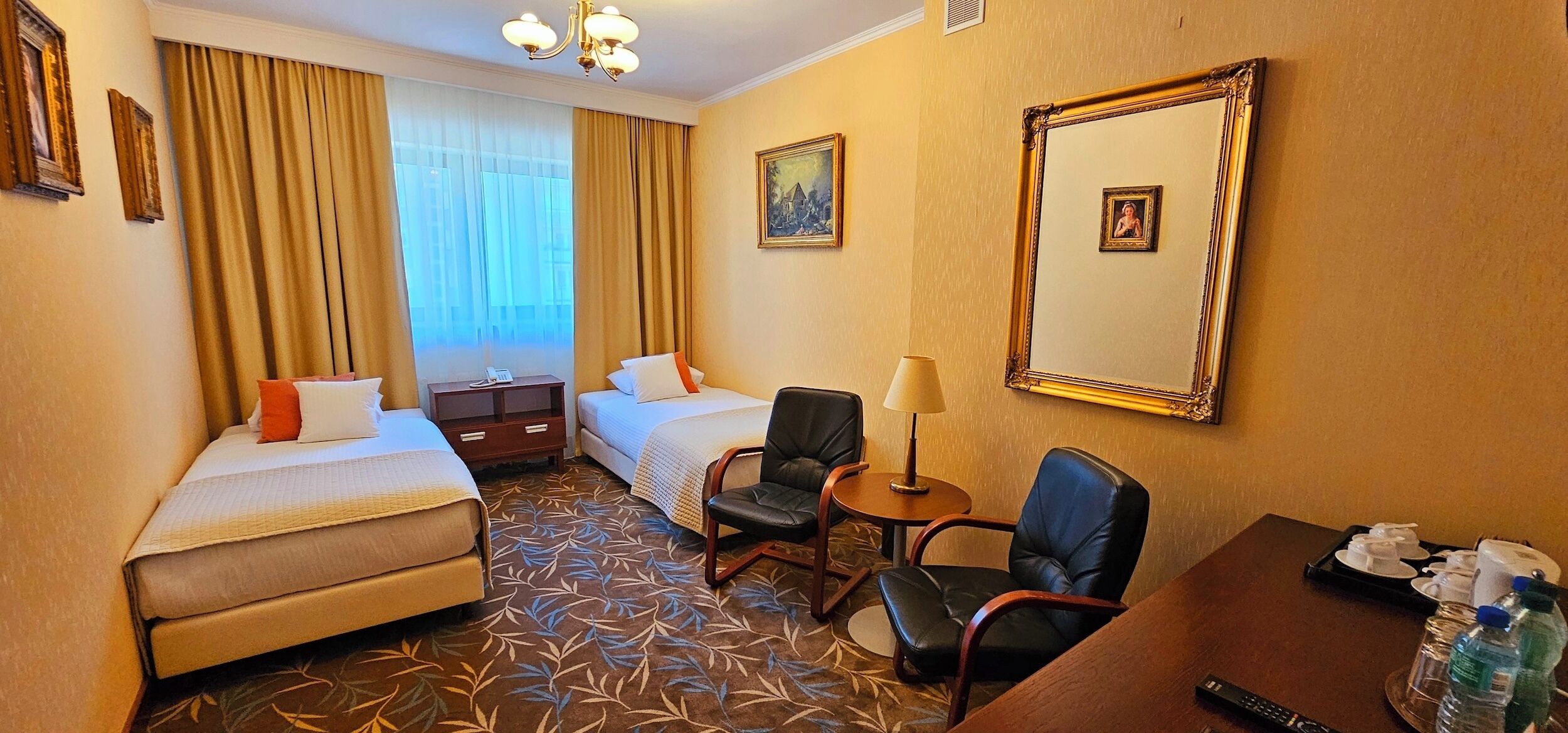 A hotel room with two beds, a desk with a lamp, a chair, a small table, and a painting on the wall.