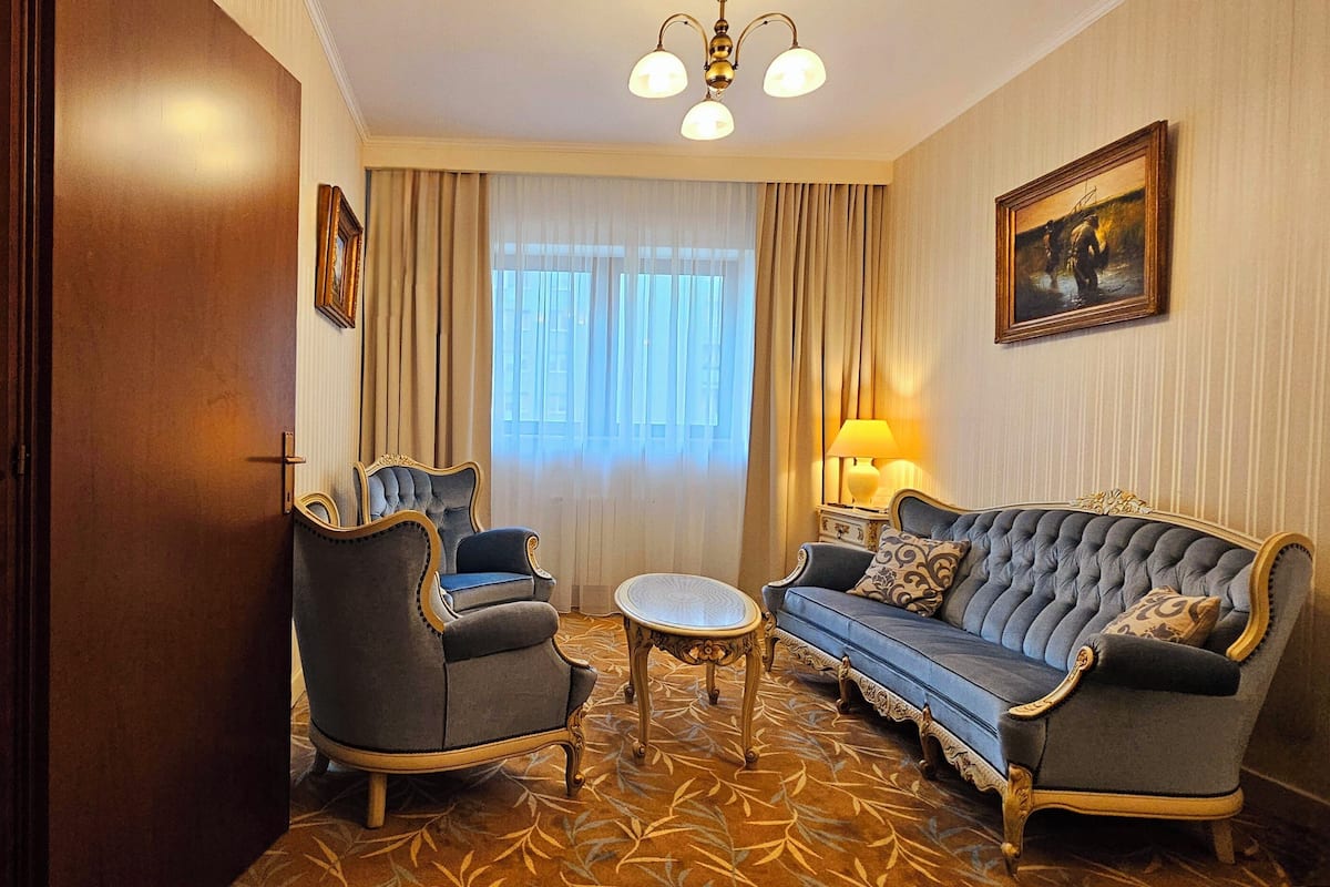 Hotellihuone, jossa on sänky, kaksi nojatuolia, sohva, pieni pöytä ja suuri ikkuna, jossa on verhot.