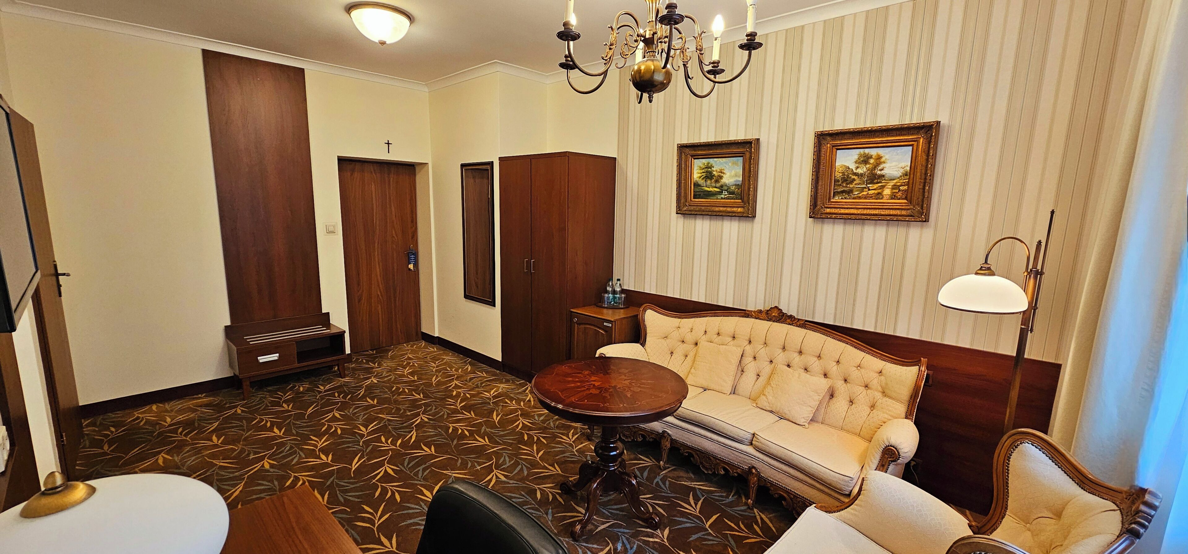A hotel room with a sofa, a round table, a chandelier, and two framed pictures on the wall.