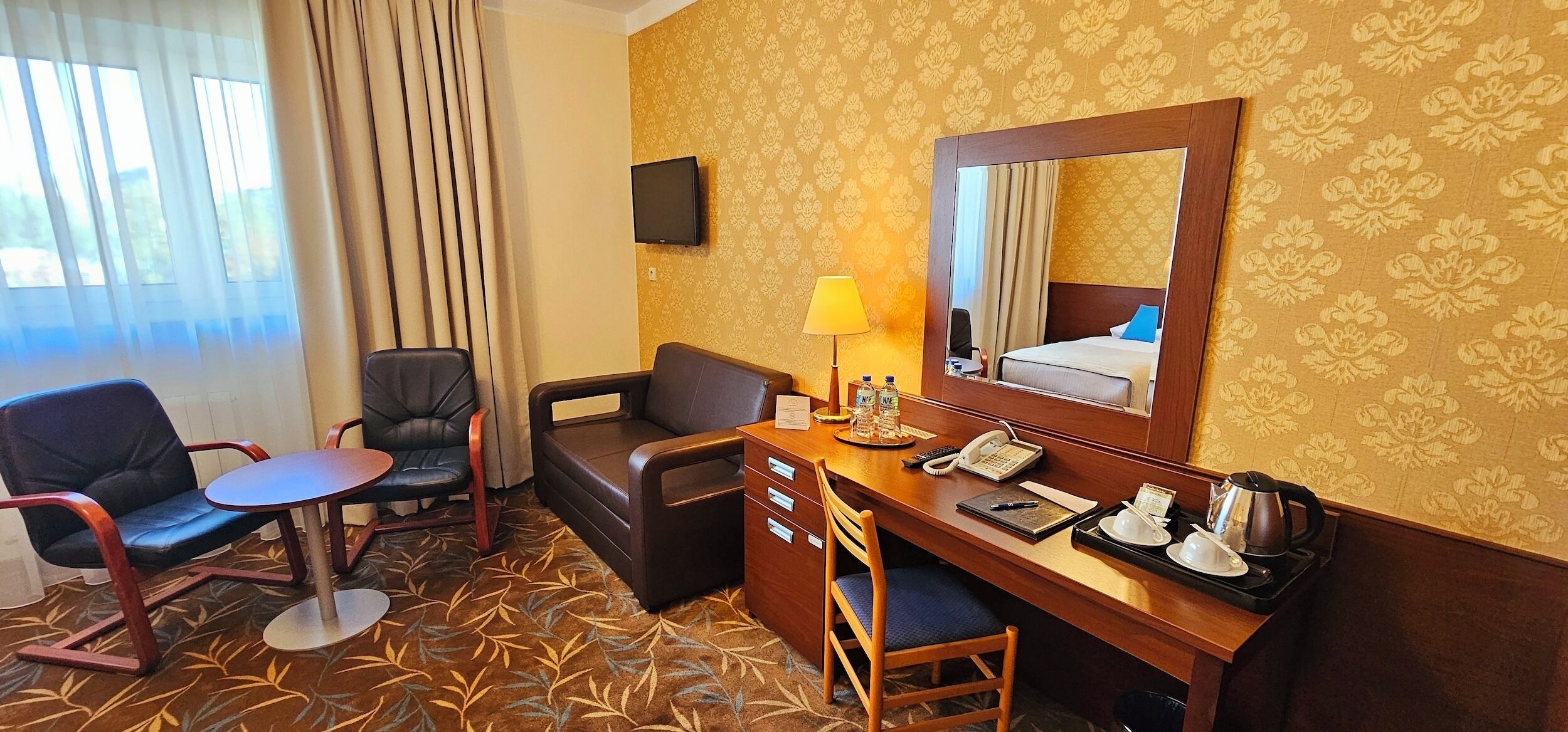 A hotel room with a desk, two chairs, a sofa, a TV, a mirror, and a window with curtains.
