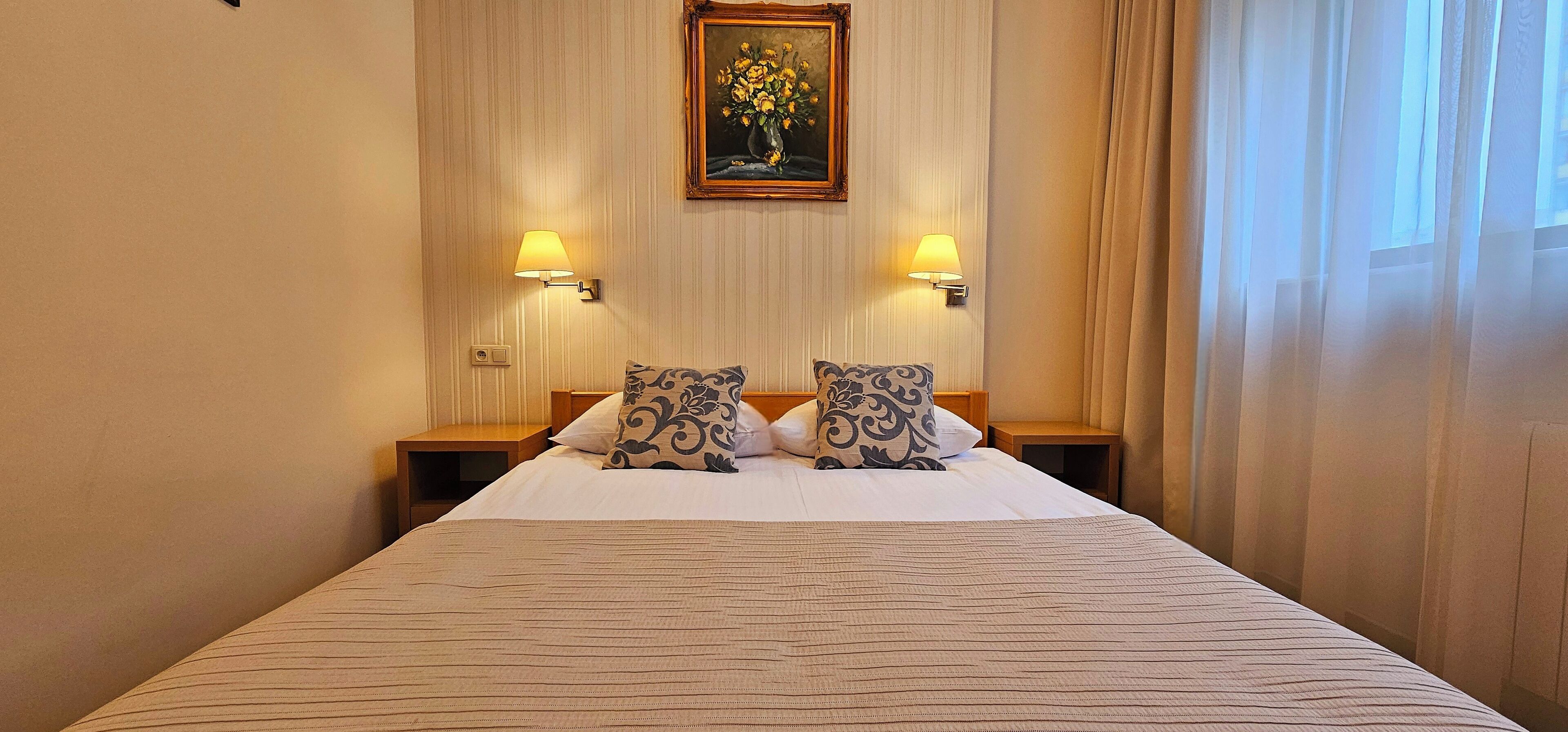 A hotel room with a large bed, two bedside lamps, a painting on the wall, and a window with curtains.