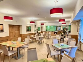 A hotel dining area with multiple tables and chairs, red pendant lights, and a kitchen in the background.