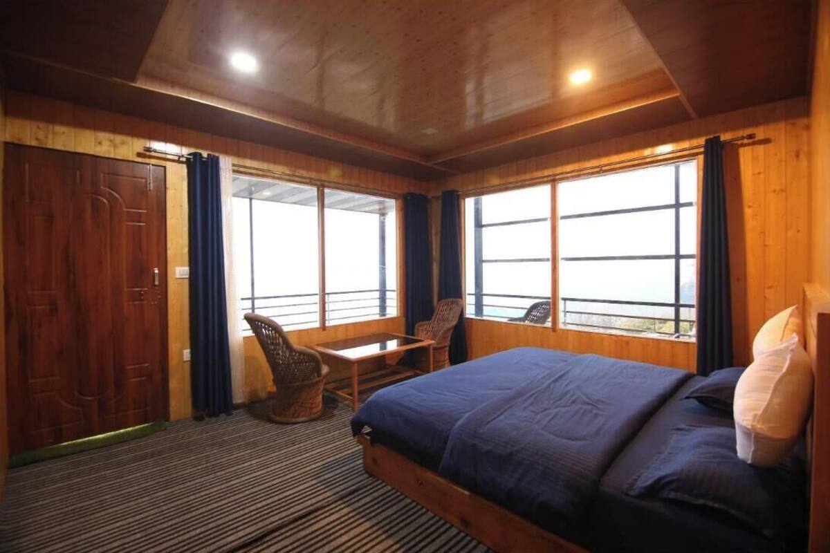 Una habitació de fusta amb un llit, dues cadires, una taula i finestres grans.
