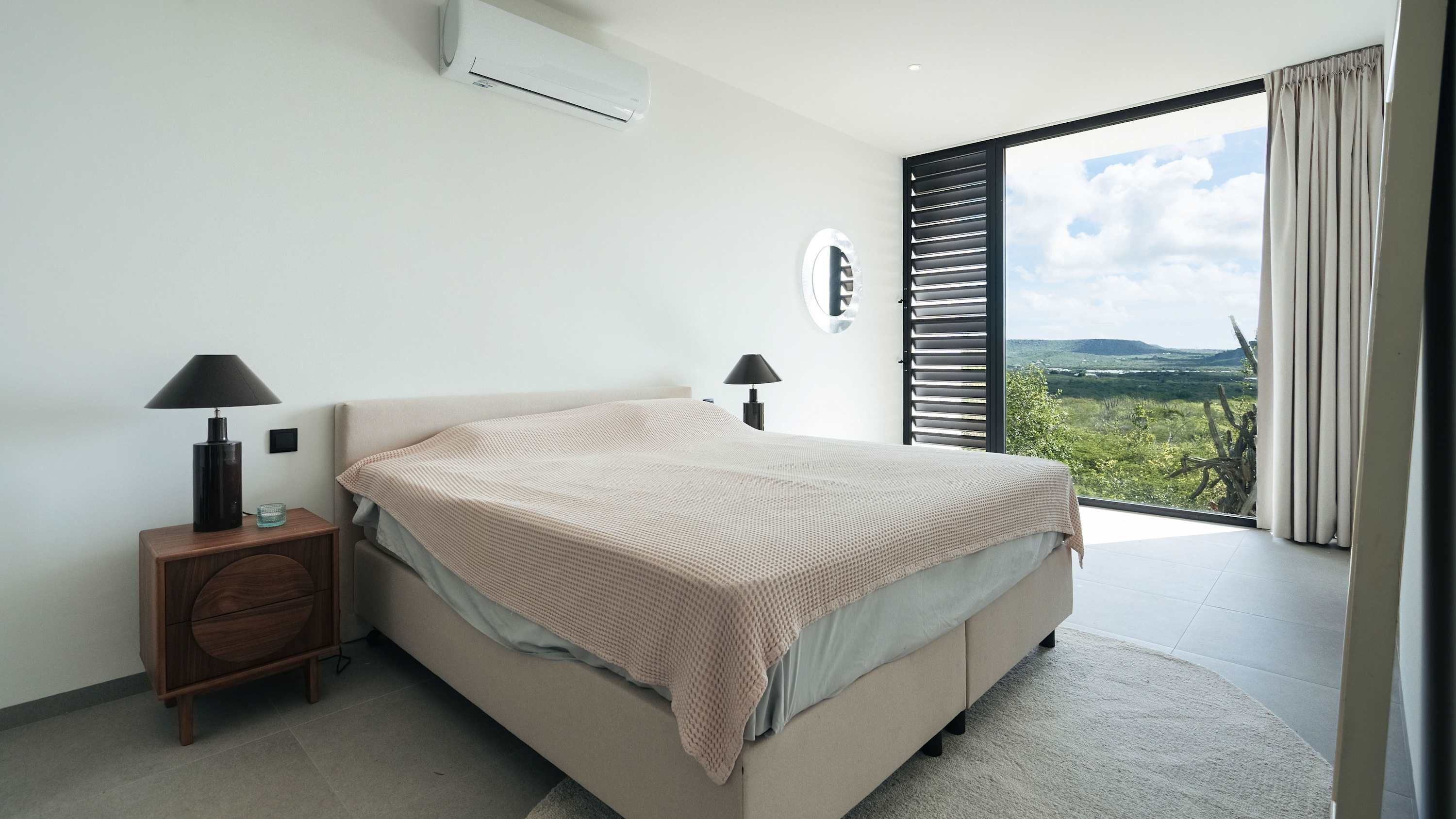 Une chambre à coucher avec un lit, deux lampes de chevet, une petite table de nuit en bois, un miroir rond et une vue sur la verdure grâce à une grande fenêtre munie de stores.