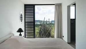 A bedroom with a bed, a nightstand, a lamp, a window with a view of a cactus and landscape, and a door leading to another room. - Villa Solara Cove Modern Design Luxury Nature (Sint Willibrordus)