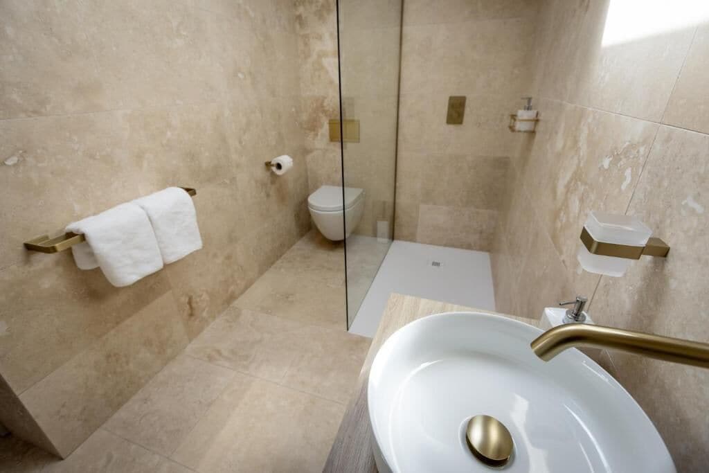 A modern bathroom with a glass-enclosed shower, a white sink with a gold faucet, and a toilet with a beige wall-mounted toilet paper holder.