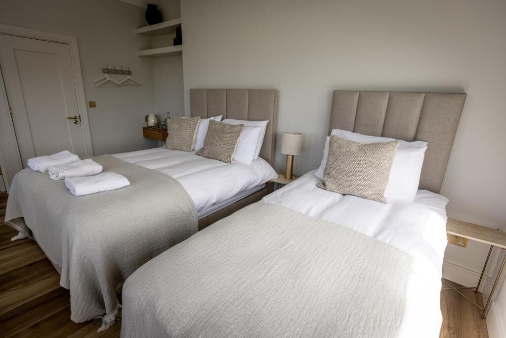 A hotel room with two beds, each with a headboard and pillows, a bedside table with a lamp, and a small shelf with items.