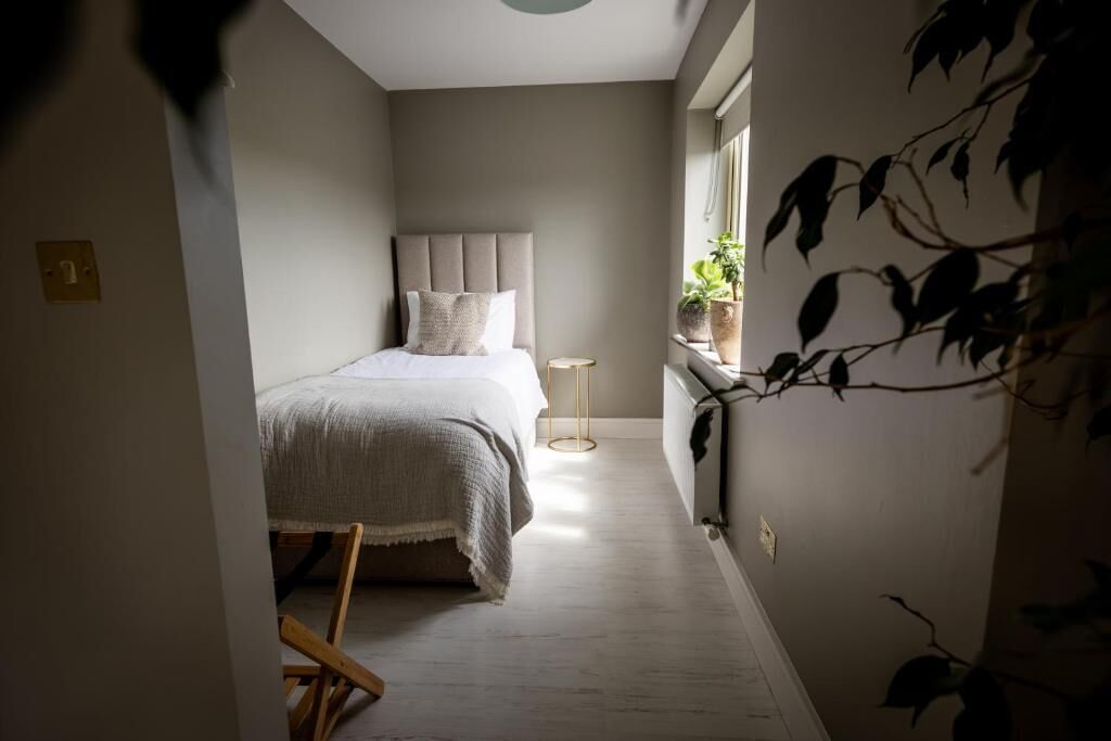 A narrow hallway leading to a bedroom with a bed, a bedside table, and a window with plants.