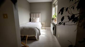 A narrow hallway leading to a bedroom with a bed, a bedside table, and a window with plants.