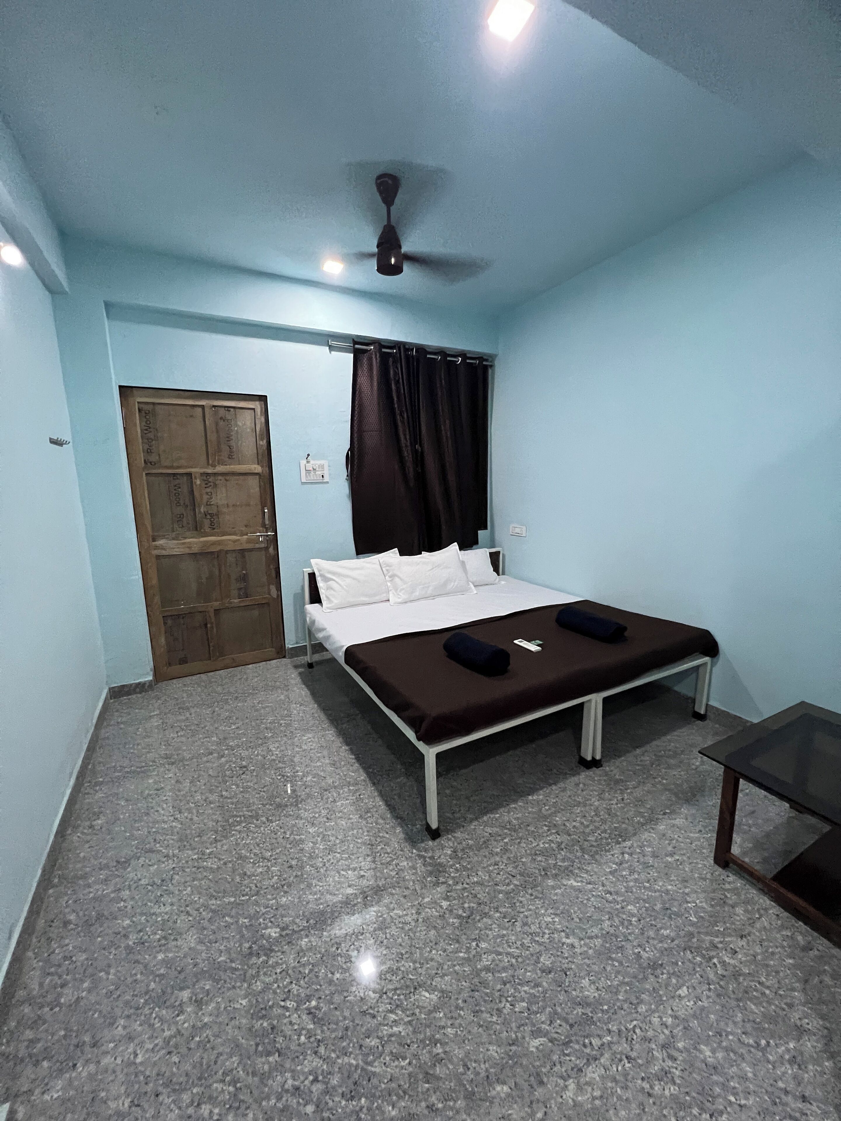 Quarto com cama, ventoinha no teto, janela com cortinas, porta e uma mesinha.