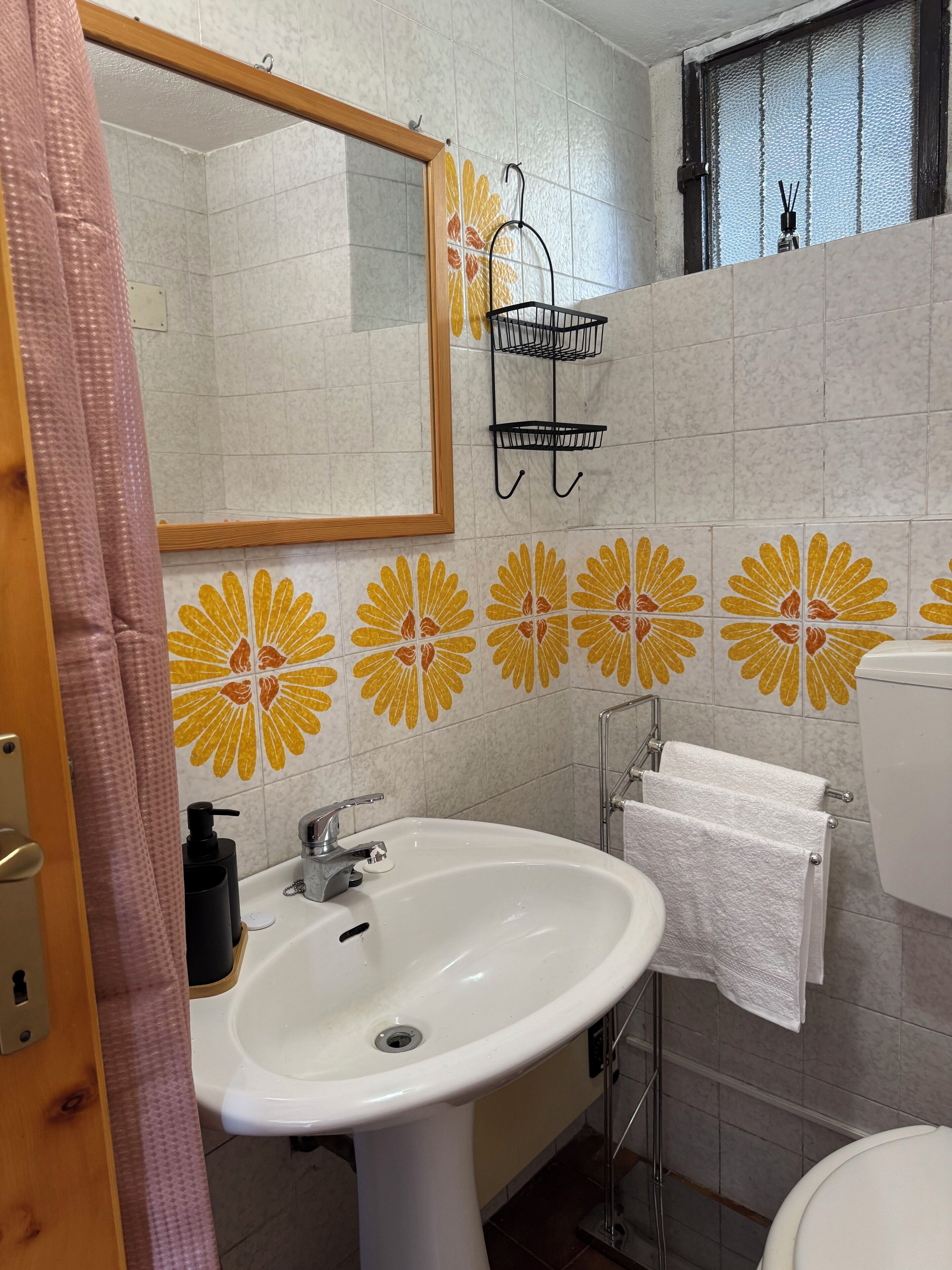 Une salle de bain avec un lavabo blanc, un miroir et des toilettes. Le mur est décoré de carreaux et il y a une fenêtre d’où l’on peut voir le ciel.