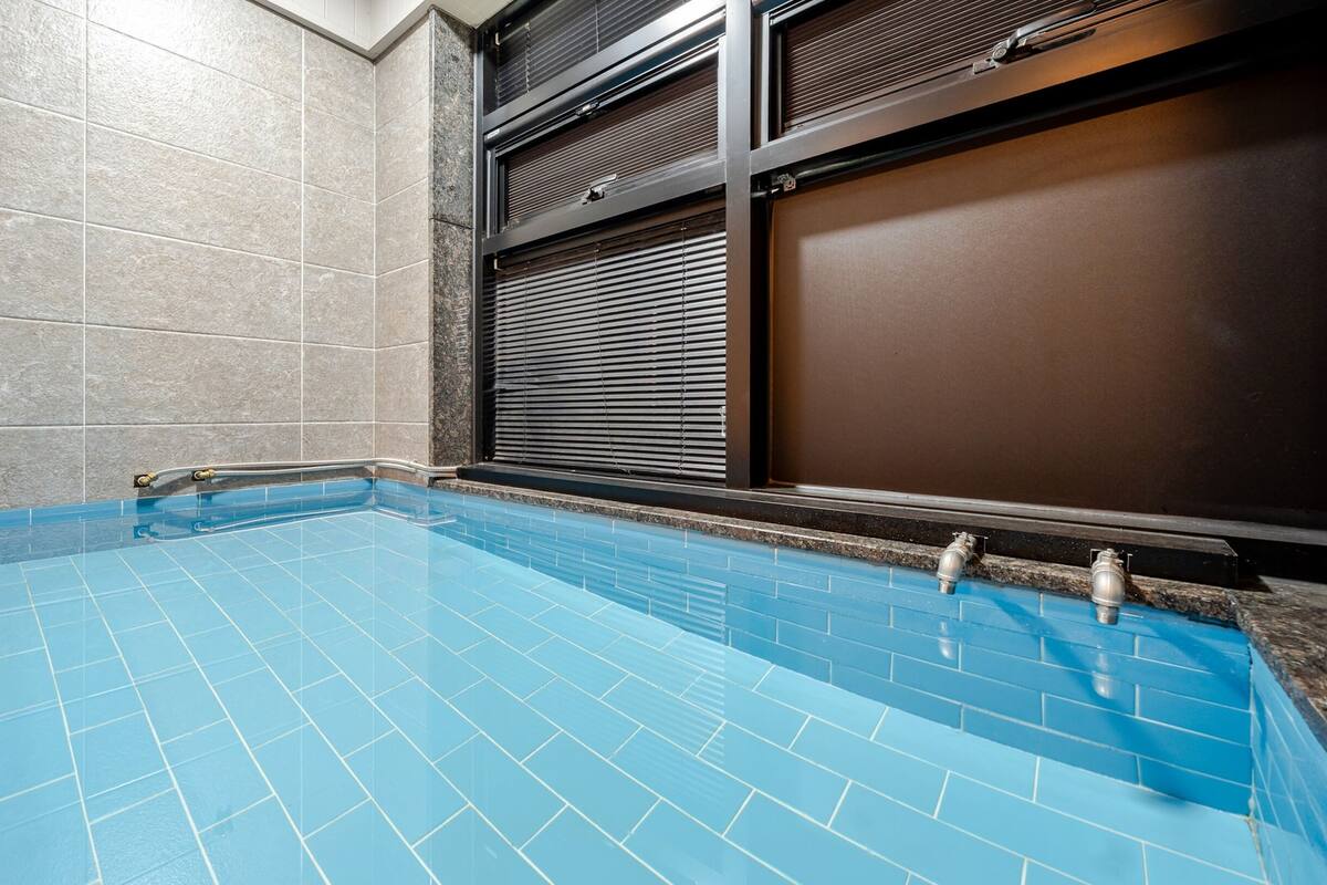 A swimming pool with blue tiles, a metal ladder, and a window with blinds.