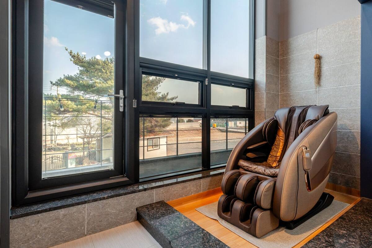 A massage chair in a room with large windows, a view of a park, and a blue wall.