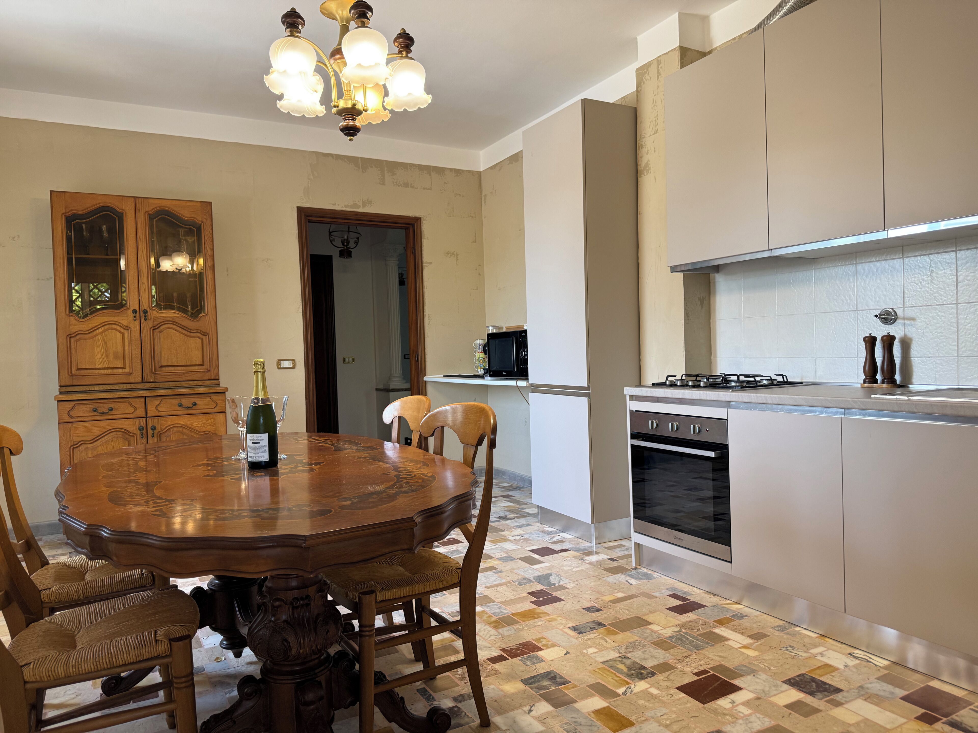 A dining area with a wooden table and chairs, a chandelier, and a kitchen with modern appliances.