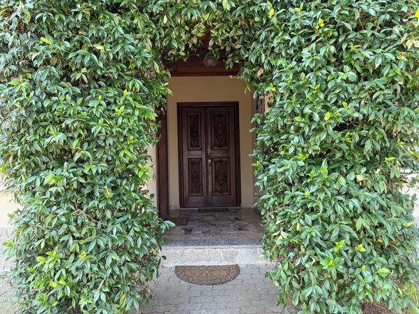A doorway framed by lush green foliage, leading to a dark wooden door with intricate carvings. - Villa With Pool Monferrato · Botto Living (Castelletto Merli)