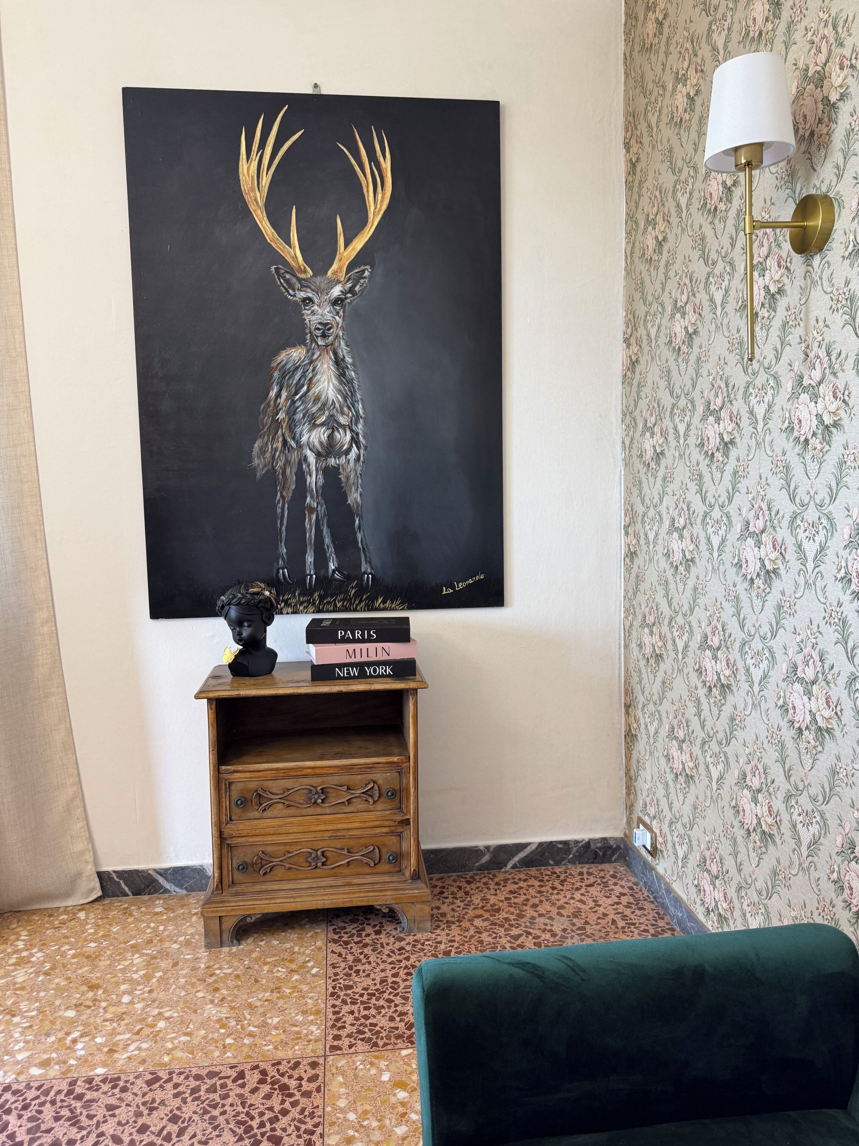 Une pièce avec un grand tableau représentant un cerf, une table d’appoint en bois avec des livres, un canapé vert et un papier peint à motifs.