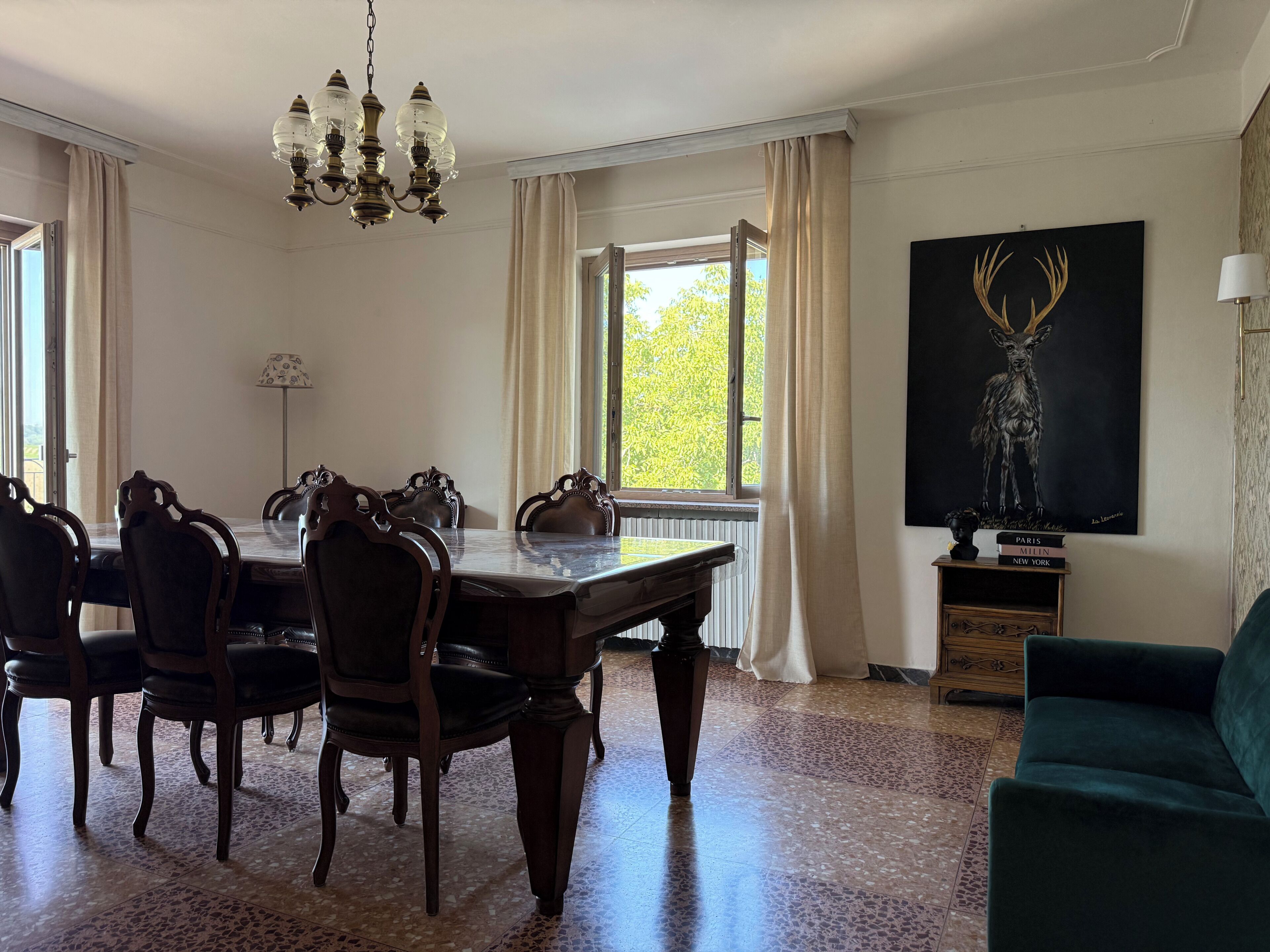 A dining area with a chandelier, a table, chairs, a window with curtains, a painting, and a lamp.