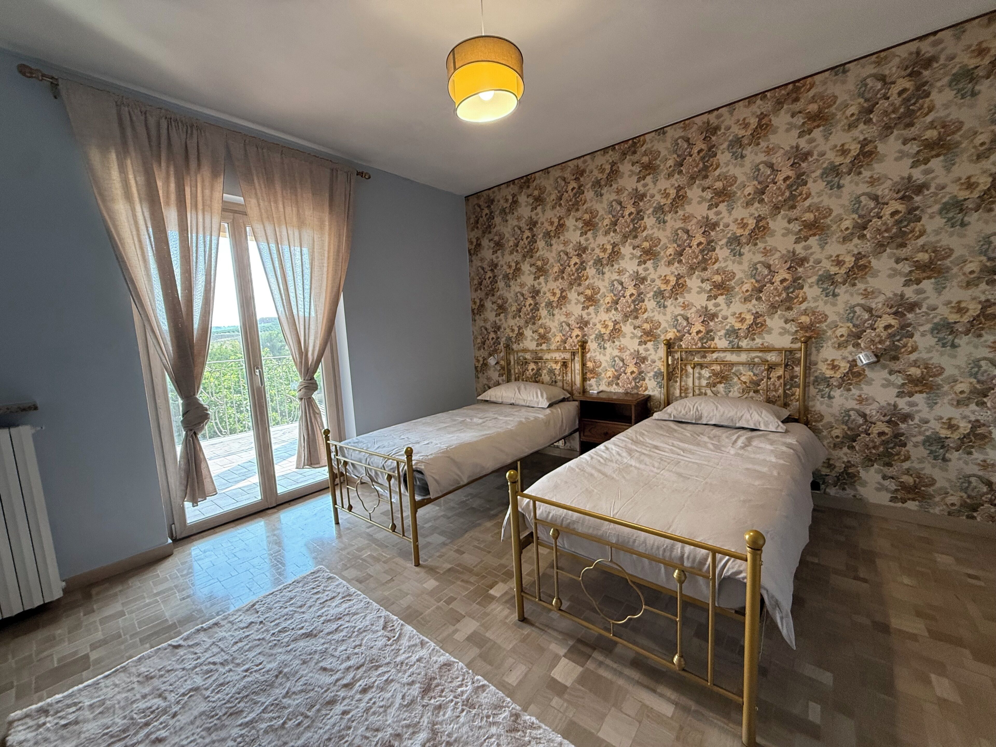 A bedroom with two beds, a radiator, a window with curtains, a wooden nightstand, and a floral wallpaper.