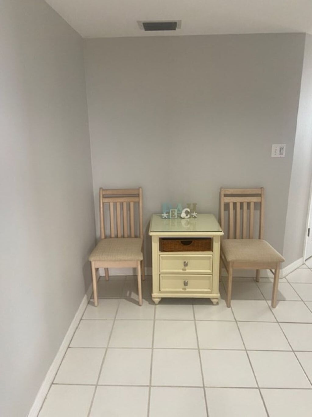 A room with two wooden chairs, a small table with drawers, and a light-colored floor.