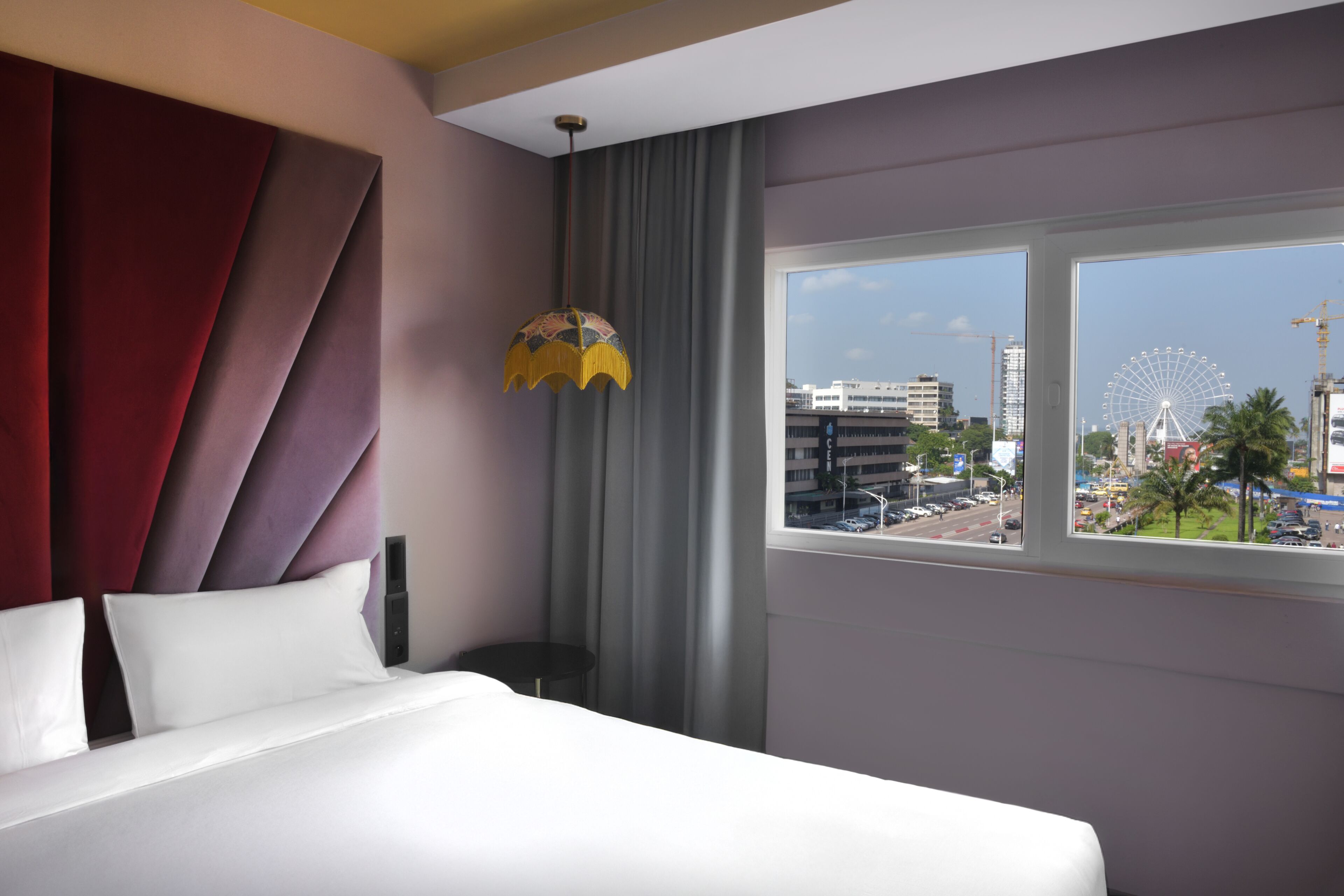 A hotel room with a bed, a view of a cityscape, a large window, and a decorative hanging light fixture.