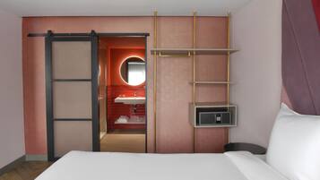Habitación de hotel con cama, puerta corredera hacia un baño, espejo redondo y estante empotrado.