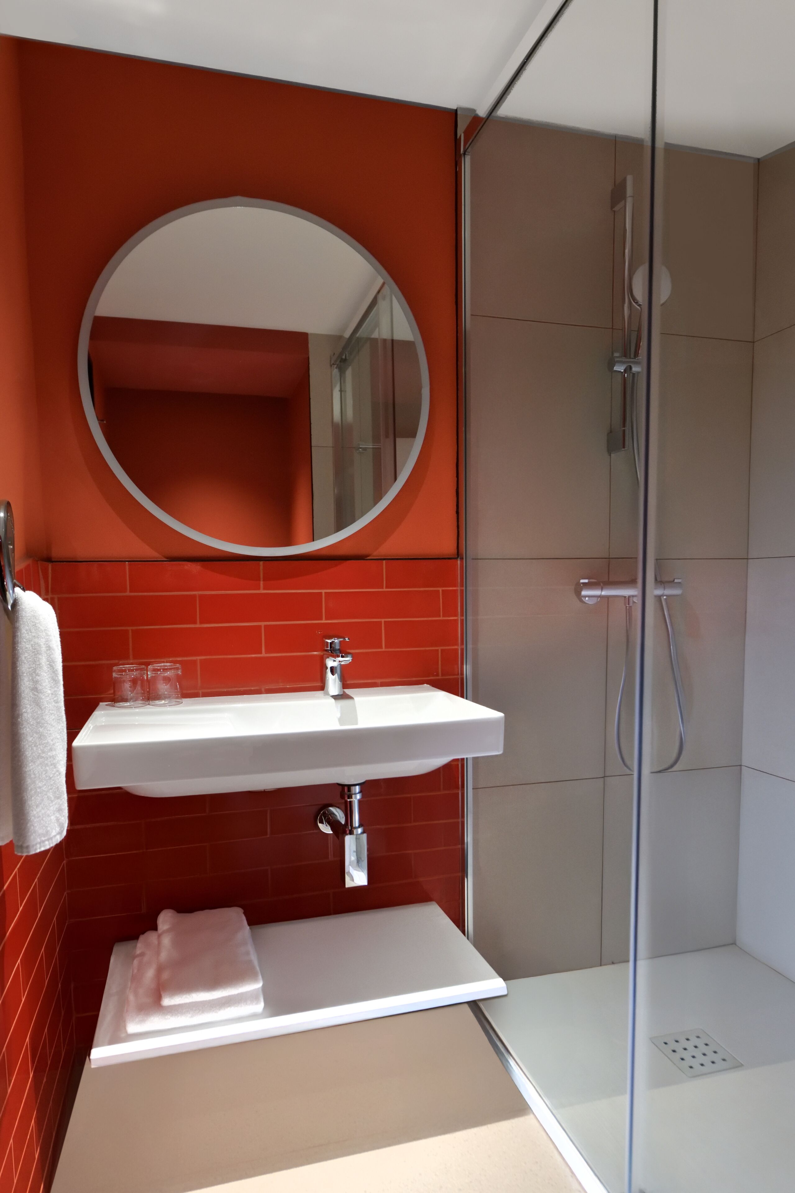 Un baño moderno con un lavamanos blanco, un espejo redondo y una ducha con mampara de vidrio.