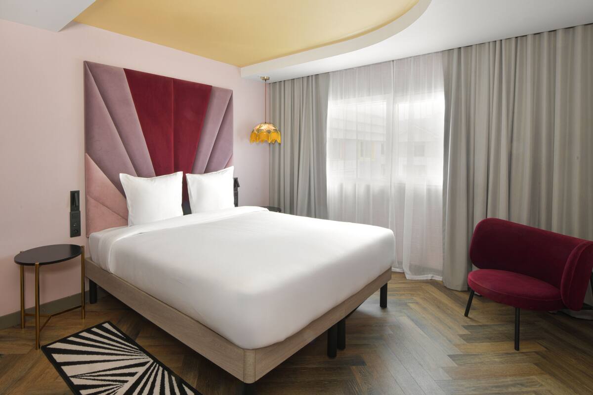 A modern hotel room with a large bed, a red chair, a small table, and a unique ceiling design.