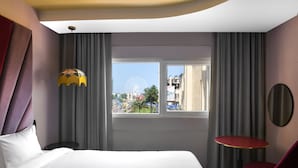 Ett hotellrum med en säng, ett fönster med utsikt över stadsbilden, en röd stol och ett litet bord.