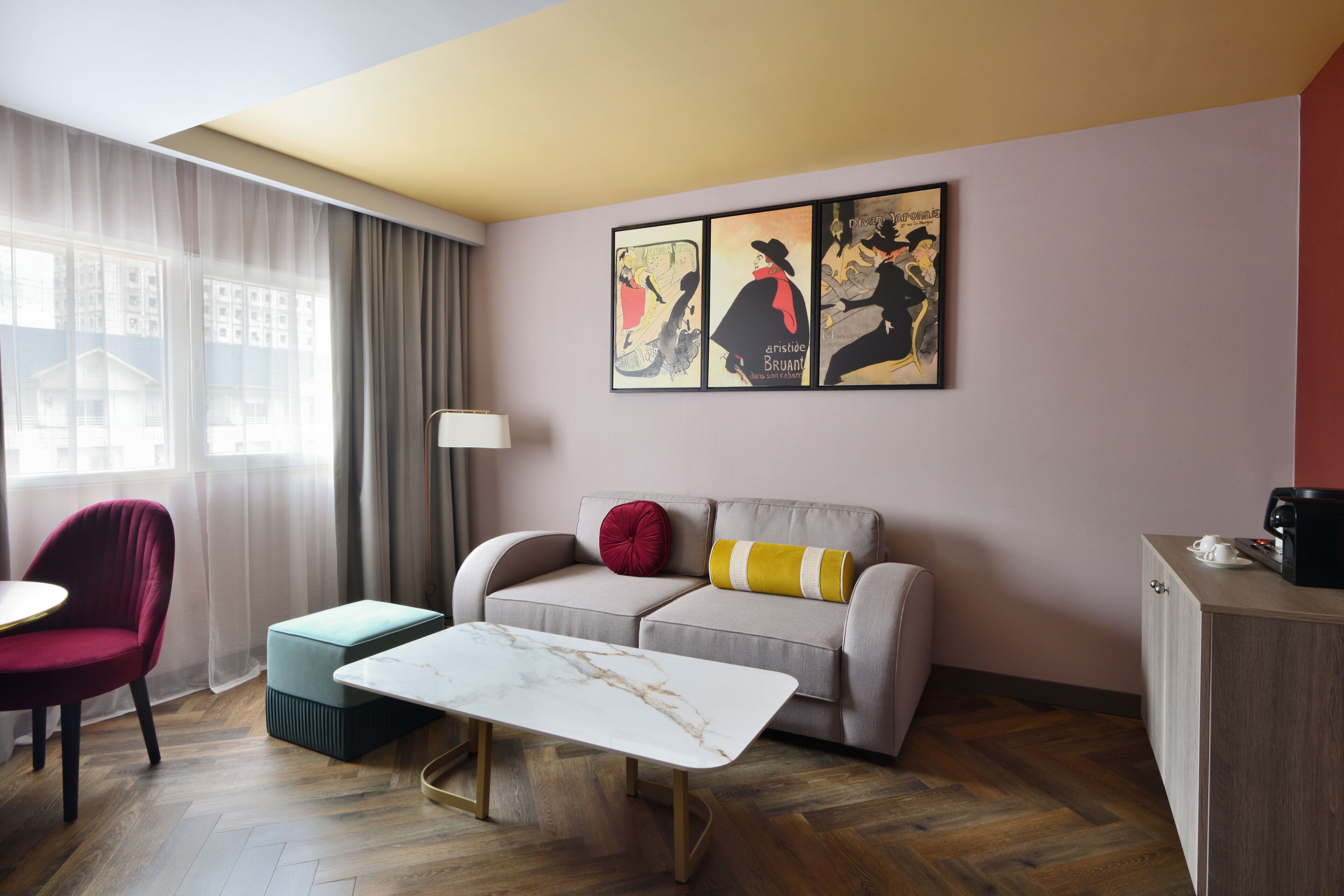 A modern hotel room with a sofa, a small table, a red chair, a cabinet, and artwork on the wall.