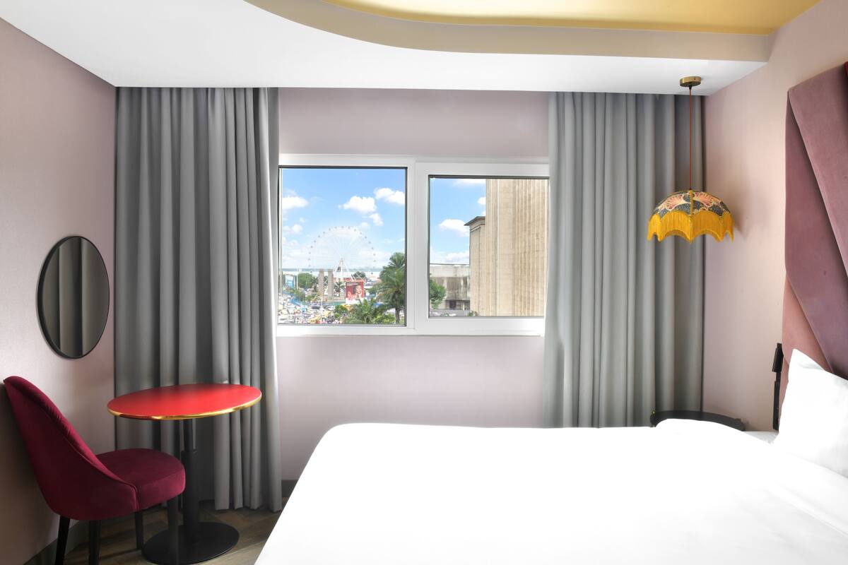 A hotel room with a bed, a red chair, a small table, a window with a view of a cityscape, and a decorative lamp.