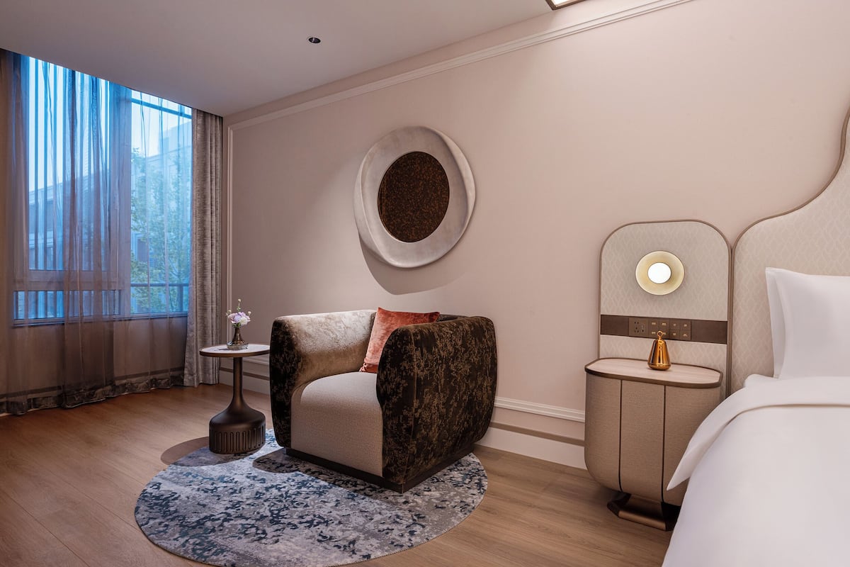 A modern hotel room with a sofa, a round rug, a small side table, a nightstand, and a bed.