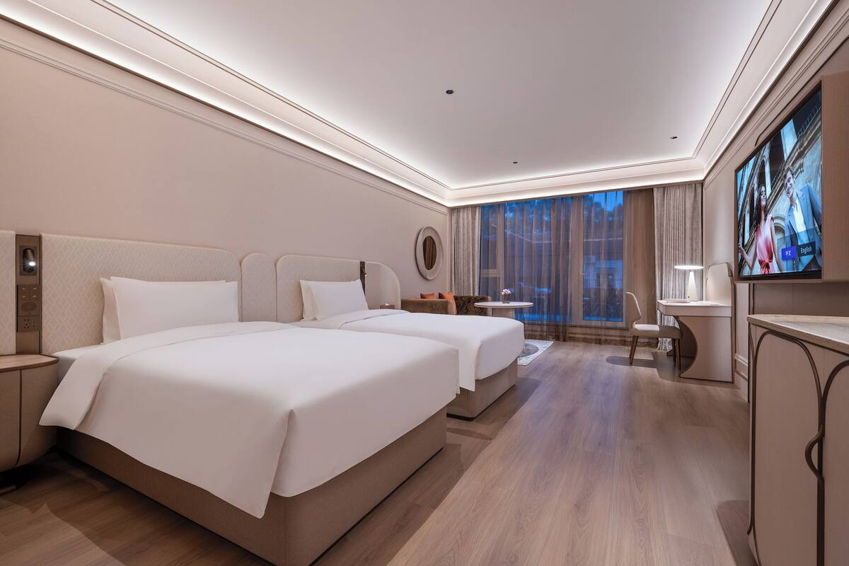 A hotel room with two beds, a TV, a desk, and a view of the city.