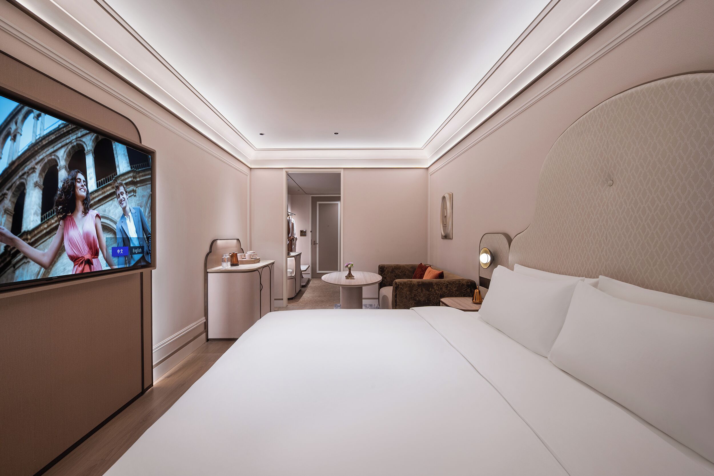 A modern hotel room with a large bed, a TV, a small round table, a sofa, and a bathroom visible through an open door.