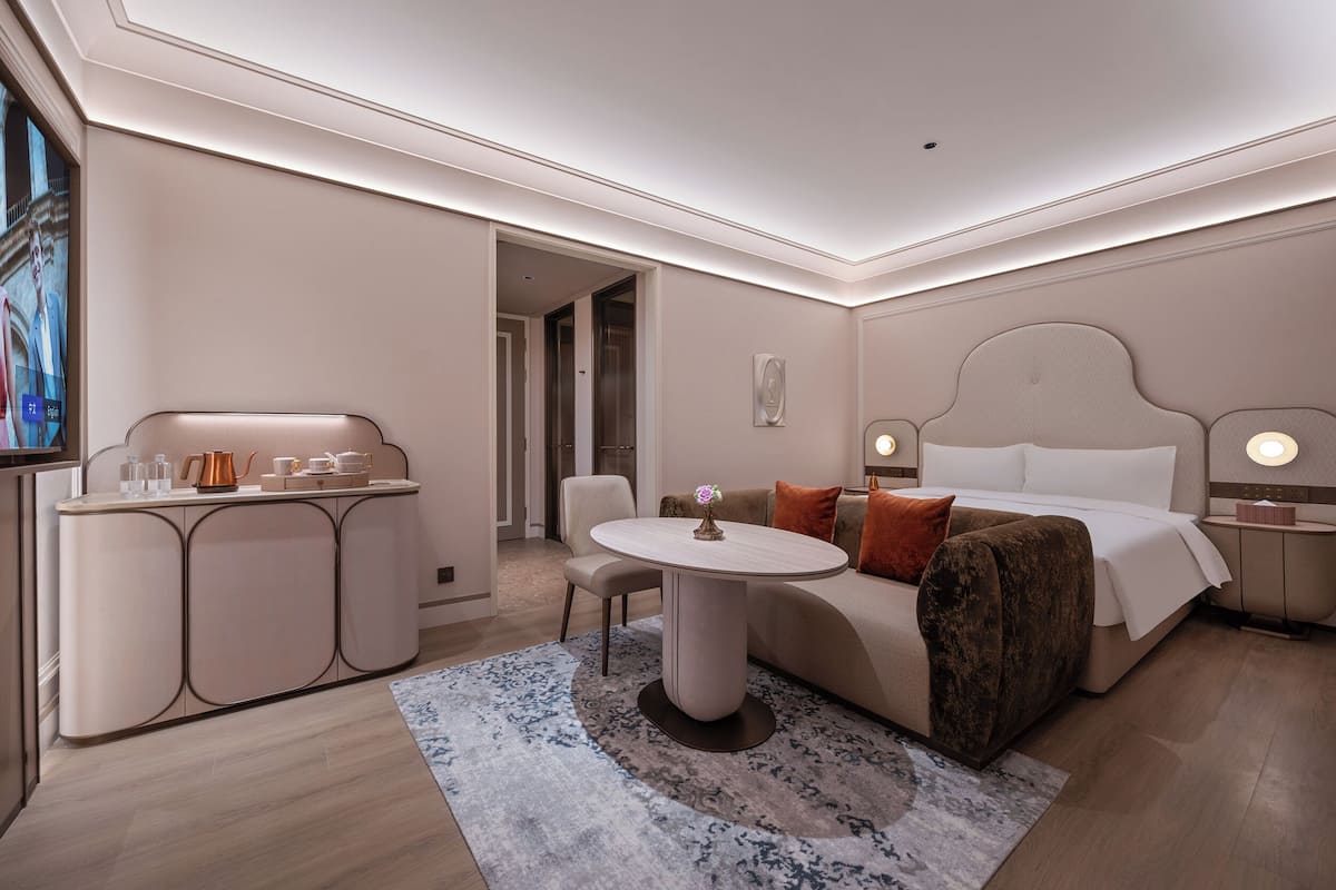 A modern hotel room with a bed, bedside tables, a round table, a sofa, and a view of the outside.