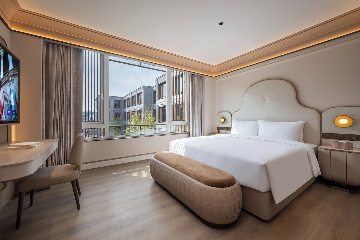 A modern hotel room with a large bed, a desk, and a view of buildings outside the window.