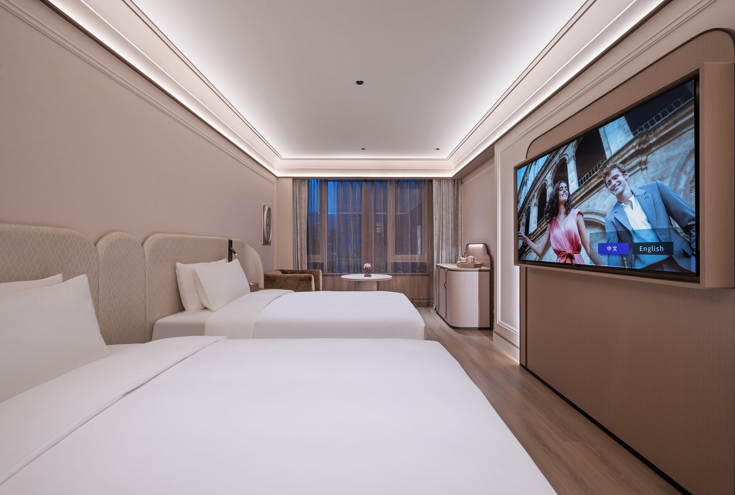A hotel room with two beds, a TV, a small table, and a window with curtains.