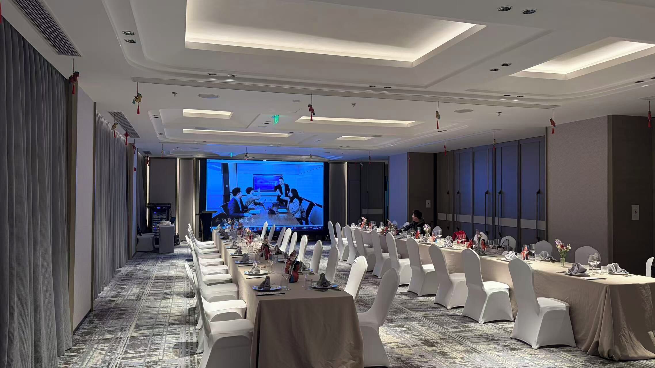 A conference room with rows of white chairs, a stage with a large screen, and a long table set for a meeting.