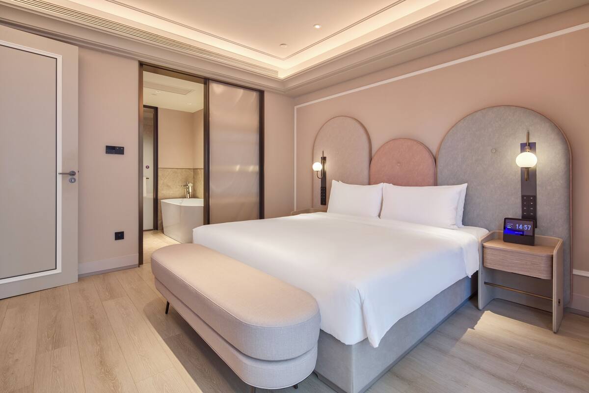 Een moderne hotelkamer met een groot bed, een bankje, een nachtkastje met een klok, een spiegel en een schuifdeur naar een badkamer.