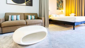A modern hotel room with a sofa, a bed, a white ottoman, and a large abstract painting on the wall. - Ibis Styles Troyes Centre (Troyes)
