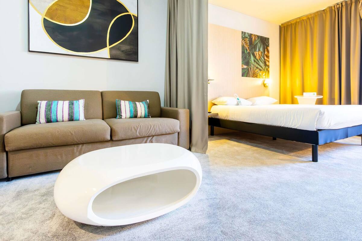 A modern hotel room with a sofa, a bed, a white ottoman, and a painting on the wall.