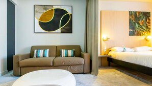 A modern hotel room with a sofa, a unique white chair, a bed, and a painting on the wall. - Ibis Styles Troyes Centre (Troyes)