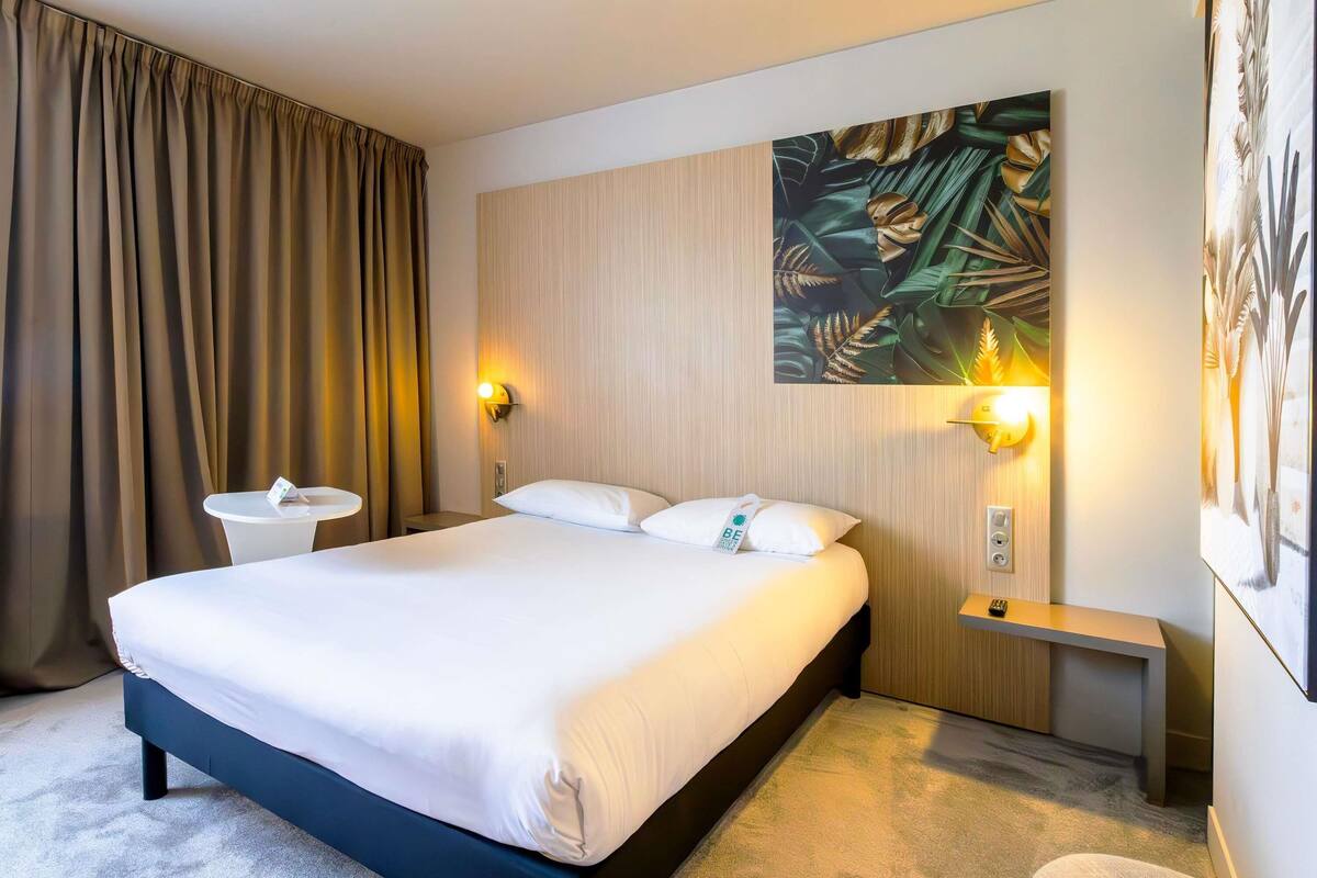 A hotel room with a large bed, two bedside tables, a wall-mounted light, and a large tropical-themed painting.