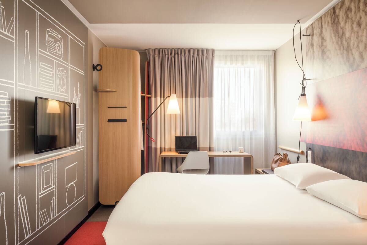 A hotel room with a bed, a desk with a laptop, a TV, and a wall with a chalkboard design.