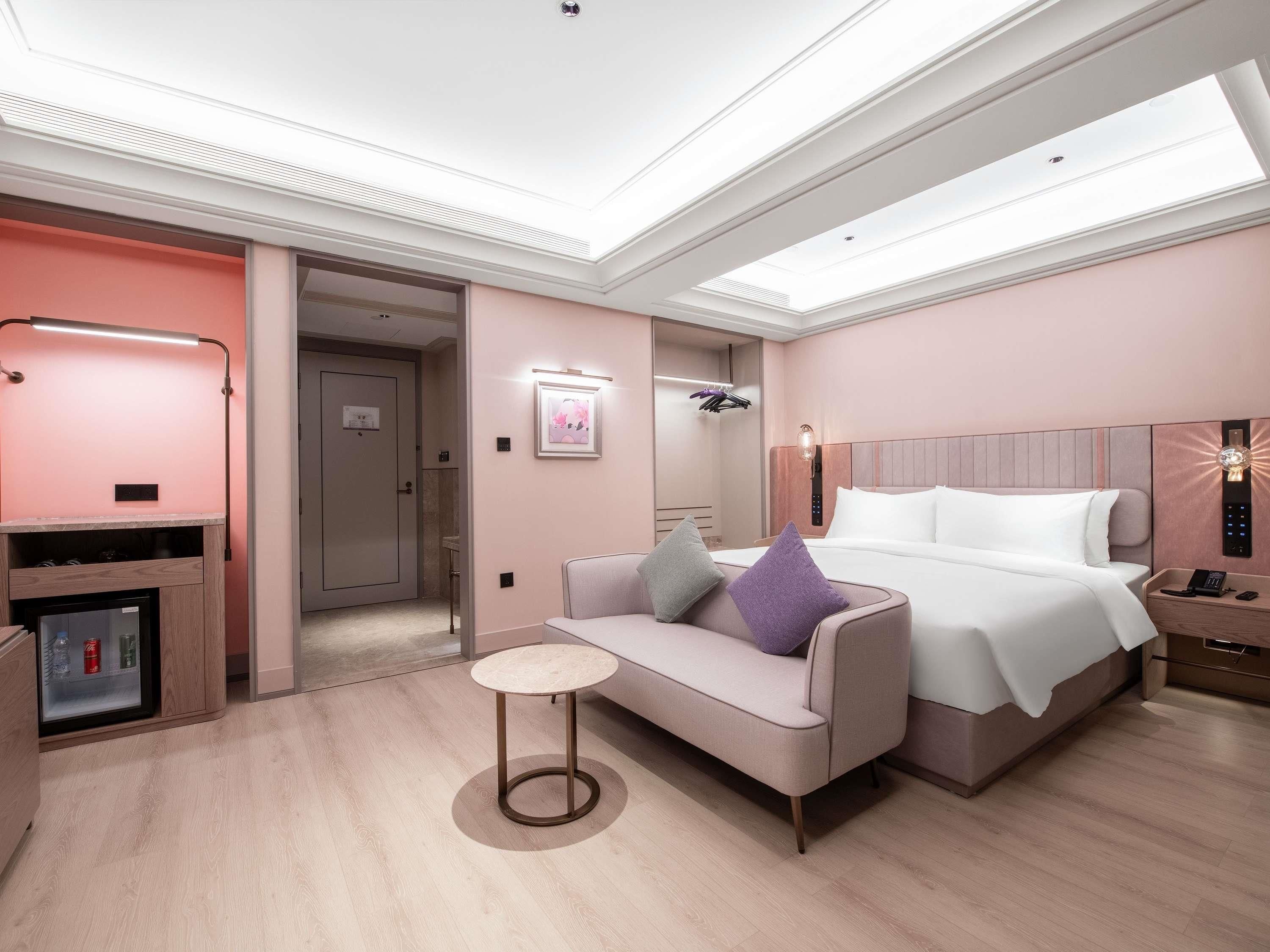 Ein modernes Hotelzimmer mit Bett, Sofa, kleinem Tisch, Minibar und einem durch eine offene Tür einsehbaren Badezimmer.
