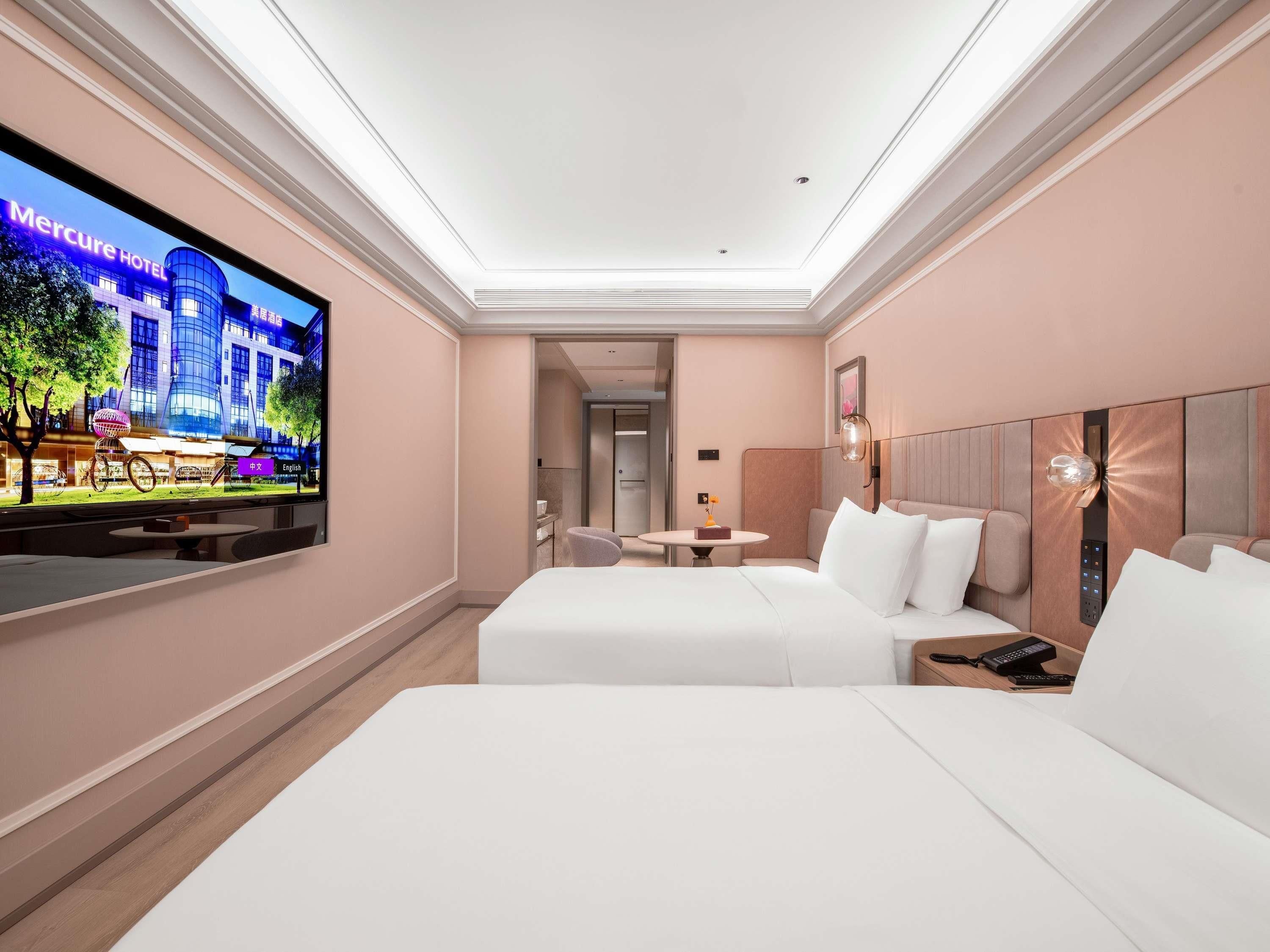 A hotel room with a large TV on the wall, a bed, a bedside table, and a small table with a chair.