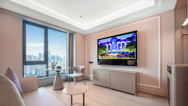 Una sala de estar moderna con un televisor grande, un sofá, una mesita y vistas a la ciudad a través de amplios ventanales.