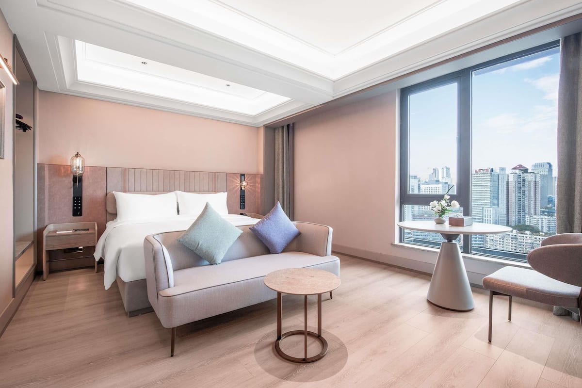 Habitación de hotel moderna con una cama grande, un sofá, una mesita redonda y vistas a la ciudad a través de amplias ventanas.