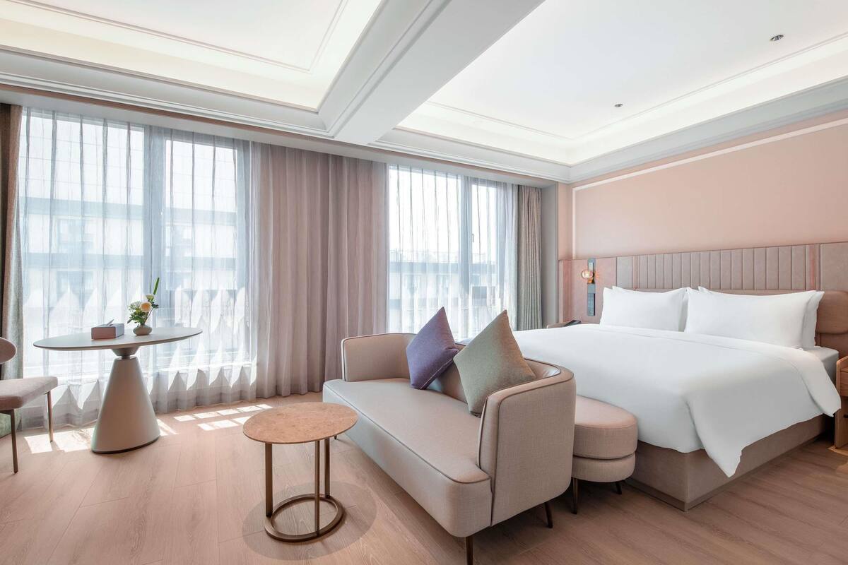 Habitación de hotel con cama, sofá, mesa redonda y vistas amplias a través de grandes ventanales.
